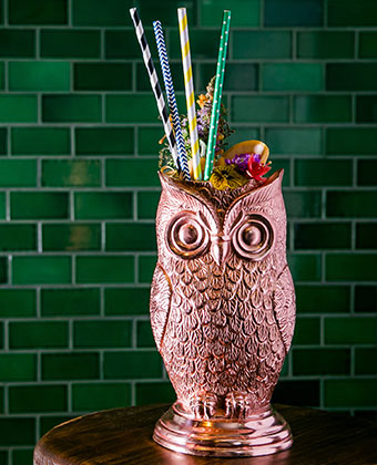 Copper Plated Owl Chalise with colorful paper straws, and flower garnish cocktail against a green brick wall